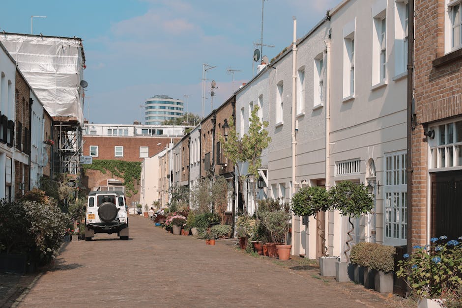 A narrow residential street with a cobbled surface, lined by terraced houses on both sides. The houses are primarily constructed with brick and painted in light, neutral tones such as white and beige, featuring white-framed windows and small front doors. Potted plants, including small trees and flowering shrubs, are arranged along the pavements in front of the buildings, adding greenery and decorative elements to the scene. On the street, a white utility van with a spare tire mounted on its rear door is parked, and construction scaffolding covered with a white tarp is visible on the left side of the image, indicating ongoing maintenance work. In the background, modern multi-storey buildings and a clear blue sky with some scattered clouds are evident, suggesting a vibrant urban environment. The scene has natural daylight, emphasizing the textures of the bricks, foliage, and cobblestone surface, and offers an observational view of a typical UK residential area that may require private waste collection or rubbish removal services from companies like House Clearance Notting Hill, which operates in this locality.