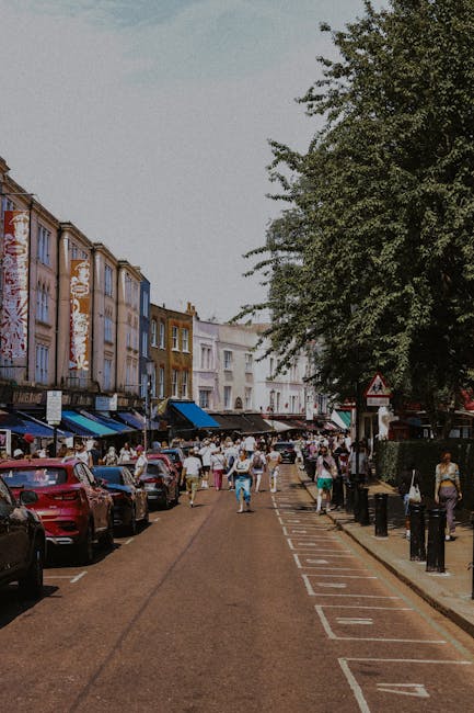 A busy street scene in an urban area during daytime, featuring a row of multi-storey buildings with colorful facades and shopfronts on the left side. Parked cars line the street along designated parking spaces, with several pedestrians walking on the pavement and crossing the road. On the right side, a large leafy tree provides shade over the sidewalk, where more people are visible. The sky appears overcast or partly cloudy, contributing to soft, neutral lighting. The setting suggests a vibrant shopping or market district, which could be served by private waste collection services for managing rubbish removal during street maintenance or clearance efforts, aligning with alternative waste handling options such as those offered by House Clearance Notting Hill.