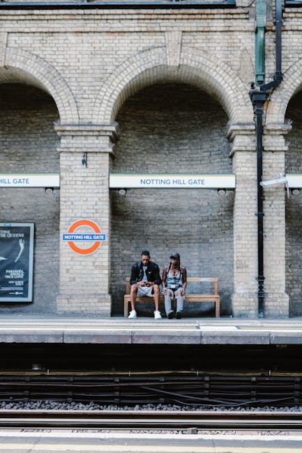 Two individuals sit on a wooden bench at the platform edge of Notting Hill Gate underground station, which features a brick façade with large arched openings. The man on the left wears a dark jacket, light trousers, and white sneakers, holding a bag on his lap, while the woman on the right wears a sleeveless top, patterned wide-leg trousers, and dark shoes, with her arms resting on her knees. Behind them, there are two white horizontal signs with blue text reading 'NOTTING HILL GATE' positioned above the arches, and a round station sign displaying the London Underground logo with the station name. To the left, a framed poster is mounted on the brick wall, partially visible. The platform surface appears clean and level, with train tracks visible in the foreground. The environment is well-lit with natural daylight, and the brickwork shows a weathered texture. This scene illustrates what could be associated with alternative methods of transport or passenger waiting areas, subtly aligning with the context of safe, private space for waste collection or disposal arrangements by companies such as House Clearance Notting Hill.