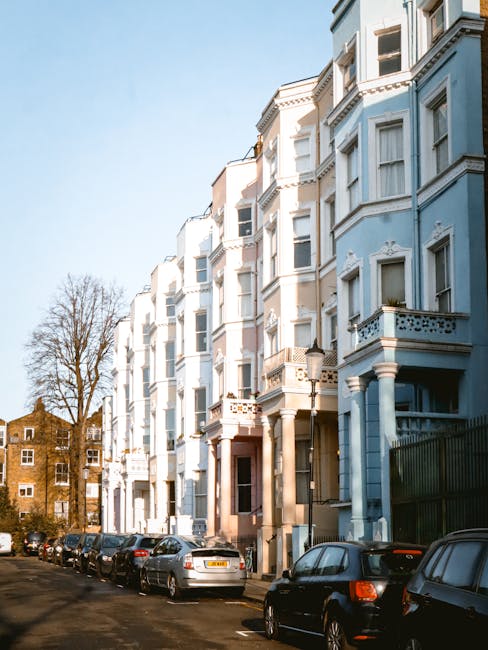 A row of multi-story residential buildings painted in light pastel shades, predominantly cream and pale blue, with detailed architectural features such as decorative cornices, bay windows, and small balconies with metal railings. The buildings are positioned along a narrow street or driveway, with several parked cars lining the curb in front, including silver, black, and dark-colored vehicles. In the background, a leafless tree is visible against a clear blue sky, indicating a winter or early spring setting. The environment appears quiet and urban, with a glimpse of other brick buildings further back. The scene relates to private property and urban living, which could contextually connect to waste management services provided by House Clearance Notting Hill for rubbish removal or building clearance, particularly at residential sites.