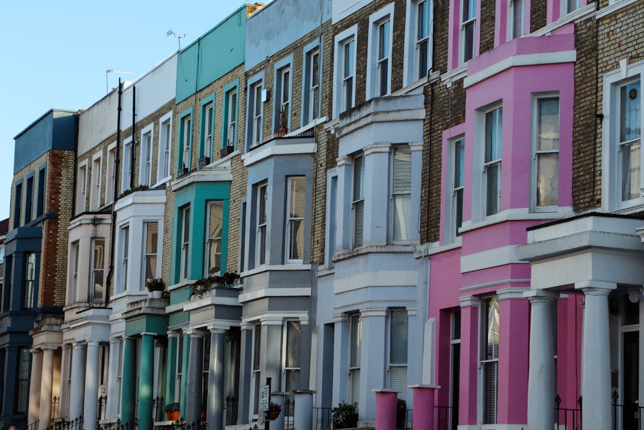 A row of Victorian-style terraced houses with ornate bay windows and decorative mouldings, painted in pastel shades of blue, white, and pink. The buildings have brick facades with some sections painted and others left as exposed brickwork. Each property features small front gardens or steps leading to the entrance doors, with black iron railings along the pavement edge. The windows are double-glazed with white frames, some open slightly, and a few have flower boxes underneath. The sky above is clear and blue, indicating daylight, and the overall scene depicts a typical residential street in an urban setting. The image subtly relates to private disposal or home clearance activities, which might involve clearing rubbish from such properties, aligning with the context of independent rubbish removal services provided by House Clearance Notting Hill.
