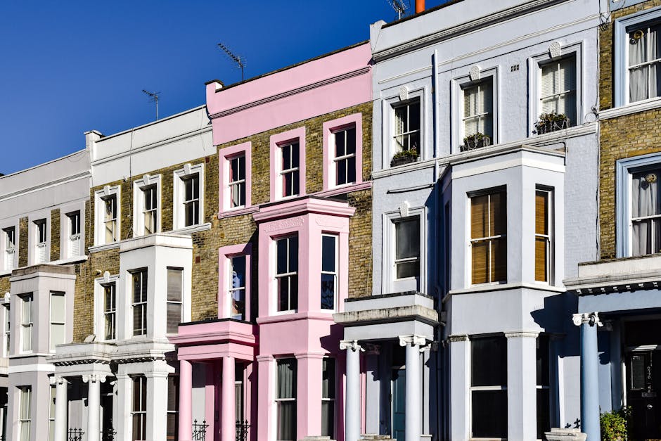 A row of Victorian-style terraced houses with three to four floors, featuring ornate brickwork and large sash windows. The buildings are painted in pastel and neutral tones, including white, light blue, and a prominent pink section in the center. Some windows have decorative flower boxes with small plants. The facades display detailed cornices and window frames, with the pink building standing out due to its bold color and architectural accents. The photograph is taken during daylight with clear blue sky, casting shadows on the buildings' fronts. The street appears clean and well-maintained, with no visible rubbish or waste materials. This setting suggests an urban residential area where private property management might involve services like waste removal or clearance, especially for general debris or damp, mouldy waste, consistent with services provided by House Clearance Notting Hill.