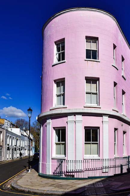 A row of Victorian-style terraced houses with three to four floors, featuring ornate brickwork and large sash windows. The buildings are painted in pastel and neutral tones, including white, light blue, and a prominent pink section in the center. Some windows have decorative flower boxes with small plants. The facades display detailed cornices and window frames, with the pink building standing out due to its bold color and architectural accents. The photograph is taken during daylight with clear blue sky, casting shadows on the buildings' fronts. The street appears clean and well-maintained, with no visible rubbish or waste materials. This setting suggests an urban residential area where private property management might involve services like waste removal or clearance, especially for general debris or damp, mouldy waste, consistent with services provided by House Clearance Notting Hill.