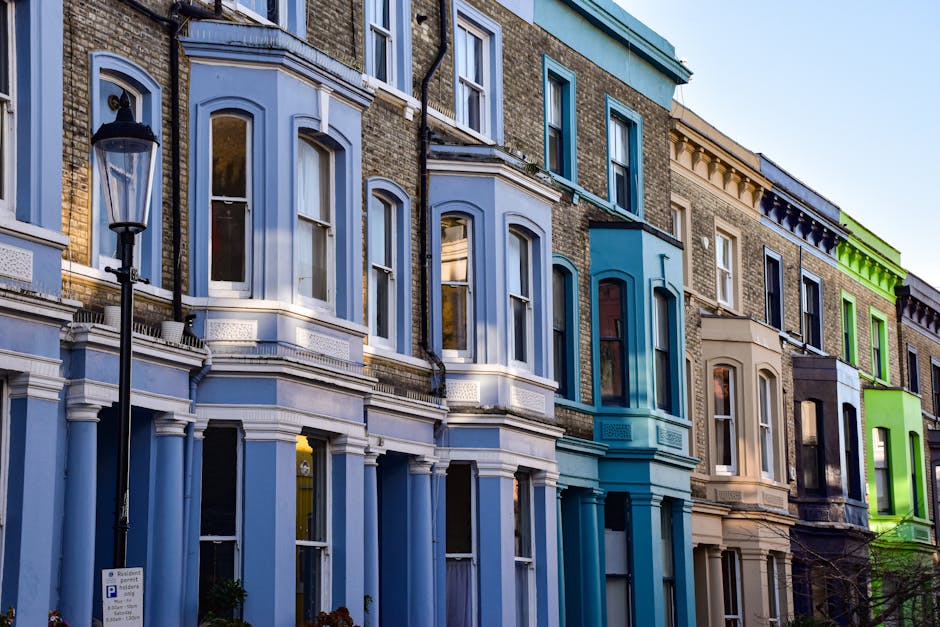 A row of Victorian-style terraced houses with multi-story brick facades, featuring large sash windows and detailed cornicing. The buildings are painted in distinct pastel shades, including light blue, beige, and bright green, with some painted accents highlighting architectural features around the windows and rooflines. In the foreground, a traditional black metal street lamp is visible, attached to a pole alongside a parking sign. The houses are situated on a quiet residential street, with the ground floor of each house showing front doors and large windows. The scene is lit by natural daylight, casting soft shadows and providing a clear view of the varied textures of brickwork and painted surfaces. The image's setting suggests an urban neighborhood where private property adaptations, such as on-site rubbish collection or scheduled clearance, may be necessary, aligning with the context of independent waste management services like those provided by House Clearance Notting Hill.