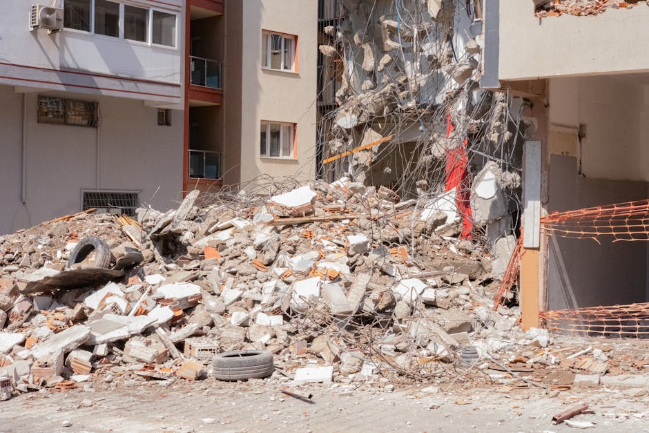 A large pile of demolition debris consisting of broken concrete, bricks, twisted metal rebar, and scattered construction materials situated in front of a multi-storey residential building with beige and white walls, orange window frames, and small balconies. The rubble extends across the ground, with some larger chunks of concrete and bent metal rods protruding upward. Among the debris, there is a discarded black rubber tire lying near the foreground on the uneven surface. To the right, a section of orange plastic safety fencing is partially visible, supported by wooden posts, indicating an active work zone or recent demolition site. The background shows the façade of the building with windows, some partially obscured by the rubble. Natural daylight illuminates the scene, highlighting the rough textures of the debris and contrasting with the smooth exterior walls of the building. This visual scene exemplifies an example where independent rubbish removal services, like those offered by House Clearance Notting Hill, may be employed to manage and clear construction waste from urban residential settings, especially after a flat or building clearance involving demolition activities.