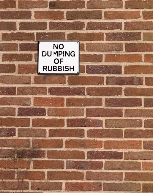 A rectangular metal sign mounted on a red brick wall displays the message 'NO DUMPING OF RUBBISH' in black capital letters on a white background. The wall consists of uniformly arranged bricks with a reddish-brown hue and visible mortar joints, extending vertically and across the entire image. The sign is positioned slightly above the center, attached with four screws, and its edges are outlined with a black border. The scene is evenly lit, with no shadows or additional objects present, suggesting an outdoor setting on a clear day. This type of signage is typically used to prevent illegal rubbish dumping in residential or commercial areas, supporting private waste management efforts by indicating that rubbish should not be left or disposed of in this location.
