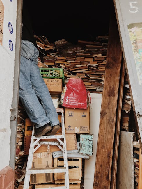 The image depicts the interior of a storage space or shed filled with a large quantity of stacked wooden planks and boards of various lengths and widths, creating a cluttered background. In the foreground, a person wearing blue jeans and black shoes is standing on a small white step ladder, reaching toward the top of the piled wood. Several cardboard boxes, some branded with the label 'Sancy,' are stacked near the person's feet, with one red plastic bag placed on top of a box. Adjacent to the boxes, a vertical wooden beam supports the structure, and a section of a wall with some posters or notices is visible on the right edge. The environment appears dimly lit, emphasizing the scale of the clutter, which could be associated with a private or on-site disposal process managed by a waste removal service such as House Clearance Notting Hill, operating within the rubbish removal category. The scene suggests ongoing clearing or sorting work, typical of a professional rubbish collection or decluttering task focused on wood and miscellaneous debris.