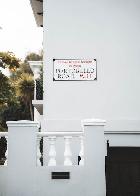 A white exterior wall of a building features a rectangular street sign mounted near the top, indicating the location as Portobello Road, with mention of the Royal Borough of Kensington and Chelsea. In the foreground, there is a low white boundary wall with decorative balusters and a black mailbox slot integrated into the wall. To the left, part of a white balcony with black wrought iron railings is visible, extending from the building. The background includes some greenery with trees and shrubs, and the scene is illuminated by natural daylight, suggesting a clear and calm environment. The overall scene represents a residential area typical of London, where private properties may require independent waste removal services, aligning with the context of alternative waste handling and rubbish clearance options in urban settings.
