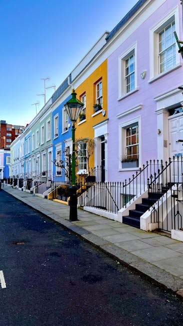 A row of colorful terraced houses on a residential street, each painted in pastel shades including lavender, yellow, and light blue. The houses feature white-framed sash windows with decorative molding, black wrought iron railings along small front steps, and black iron wall-mounted lanterns. A traditional black lamppost stands on the sidewalk in front of the houses. The street is paved with asphalt, with a narrow concrete sidewalk lined with gray paving slabs. Some houses have small front gardens or steps leading up to white front doors, with flower boxes beneath the windows. The scene is illuminated by natural daylight, with a clear blue sky visible above. House Clearance Notting Hill occasionally performs on-site clearance or private waste handling, which may include collecting bulky waste such as discarded furniture or household items, in environments like this residential street, supporting alternative waste disposal options outside of local authority collections.