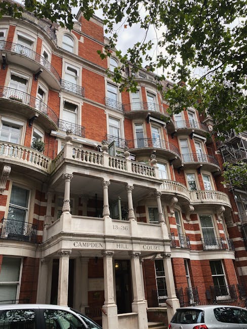 A multi-story residential building with a classic brick façade and decorative white stone balcony railings. The building features rounded corner sections with projecting bay windows on each floor. The entrance area, supported by columns, displays an inscription reading 'Campden Hill Court' and the numbers '13-36'. The front of the building is set against a street with parked cars visible at the bottom of the image. Overhanging green tree branches partially obscure the upper part of the building, and natural daylight highlights the textures of the red brickwork and the cream-colored stone details. The scene suggests an urban environment with well-maintained architectural features typical of London residential blocks, and the image may relate to the context of on-site clearance or private waste management by companies like House Clearance Notting Hill, specializing in rubbish removal services in the area.