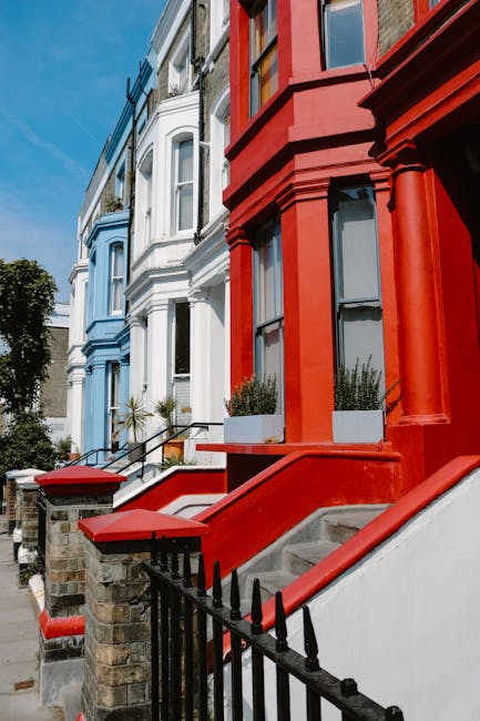 The image depicts a row of Victorian-style terraced houses situated on a residential street with a clear blue sky overhead. The buildings are painted in distinct colours: the first house on the left is painted in a light blue with white window frames and decorative detailing; the adjacent house is predominantly white with ornate mouldings and a bay window. On the right, a red-painted house features large, rectangular windows with black frames and decorative columns supporting the façade. In front of the red house, a small white concrete staircase with metal railing leads up to the entrance, accompanied by planter boxes filled with green plants. The pavement is paved with concrete slabs, and a short brick wall with black iron fencing runs alongside the street, separating the houses from the sidewalk. The scene lacks any visible rubbish or waste, reflecting a clean and well-maintained urban environment. House Clearance Notting Hill occasionally provides private on-site clearance services suitable for homeowners undertaking estate or house clearances in the area, supporting alternative waste handling for property refurbishment or relocation.