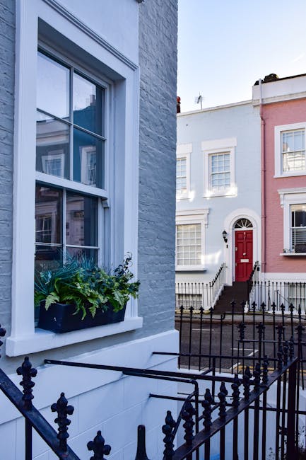 The image shows the exterior of a row of terraced houses with distinct architectural features. In the foreground, there is a white-painted brick wall with a large, double-hung sash window framed in white, featuring multiple panes separated by white muntins. A window box with green leafy plants and foliage sits below the window, adding a touch of natural greenery. Adjacent to the brick wall, a black wrought iron fence with decorative finials runs along the front of the houses, enclosing a small front yard area. In the background, three additional terraced houses are visible, each with a different colour scheme: one is painted in a light pastel pink, with a white decorative cornice and sash windows, and another in a white facade with matching window frames. The house with the pink facade features a prominent red door accessed via a short flight of black steps with matching white risers, flanked by black metal railings. The overall scene is illuminated by natural daylight, creating a clear and neutral atmosphere that emphasizes the architectural details and the urban setting, which is indicative of a typical residential street suitable for private waste collection or on-site clearance services by companies like House Clearance Notting Hill.