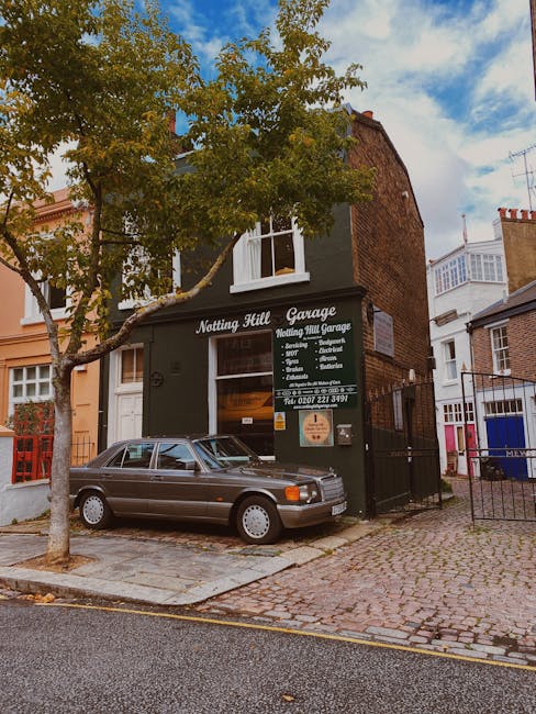 A two-storey brick building with a dark green exterior, housing the Notting Hill Garage, features a large sign displaying services such as vehicle servicing, repairs, and MOT testing. The building's ground floor has a glass window and a door, both partially covered by the sign, with the garage's name painted on the facade. In front of the garage, a silver-grey four-door sedan is parked on a paved street, with a small, leafy tree on the left side providing partial shade. The surrounding area includes neighboring buildings with brick and stucco facades, some with visible windows and balconies, set along a cobbled roadway with a concrete curb. The scene is illuminated by daytime natural light, with a blue sky overhead and scattered clouds, indicating a clear, bright day. This setting illustrates a typical urban environment where private and independent rubbish collection services, such as those offered by House Clearance Notting Hill, may be employed for specialized waste management needs, especially for commercial or on-site clearance scenarios.