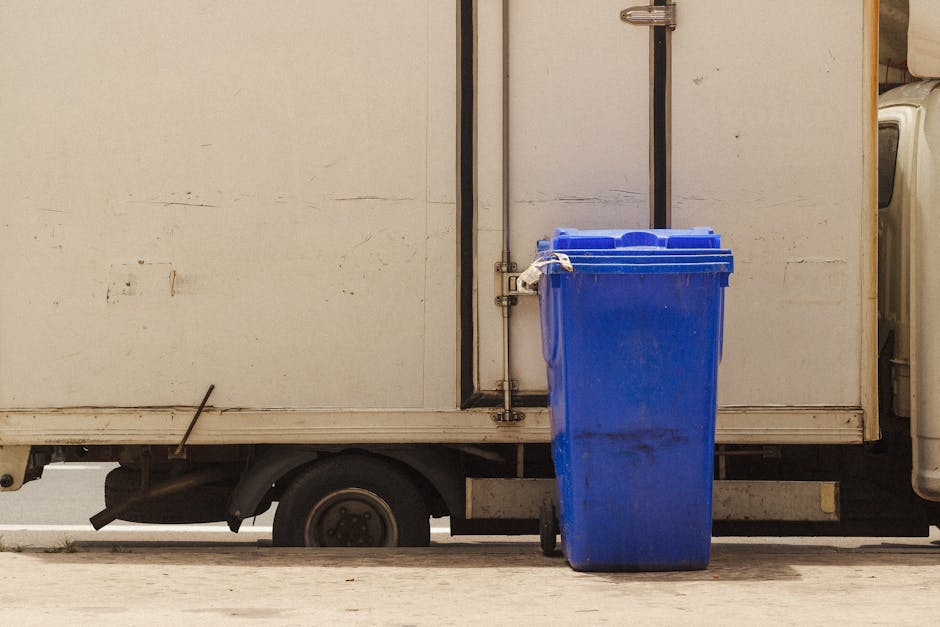 A blue wheeled rubbish bin made of durable plastic with a textured surface and a slightly scratched finish, positioned upright on a concrete pavement next to a small white delivery truck. The bin is placed close to the back of the truck, with its lid closed, and appears to contain waste or recyclables. The truck's side panel is plain, showing signs of dirt and minor wear, and is framed by a metal strip near the door. The scene is lit with natural daylight, casting soft shadows, and the setting suggests an area used for waste collection, possibly for private disposal services like those offered by House Clearance Notting Hill. The environment indicates an urban or residential street where waste is independently gathered and prepared for removal, aligning with the context of alternative rubbish collection services in the area.