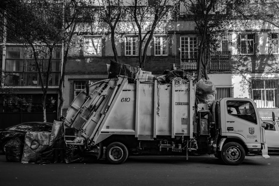 A blue wheeled rubbish bin made of durable plastic with a textured surface and a slightly scratched finish, positioned upright on a concrete pavement next to a small white delivery truck. The bin is placed close to the back of the truck, with its lid closed, and appears to contain waste or recyclables. The truck's side panel is plain, showing signs of dirt and minor wear, and is framed by a metal strip near the door. The scene is lit with natural daylight, casting soft shadows, and the setting suggests an area used for waste collection, possibly for private disposal services like those offered by House Clearance Notting Hill. The environment indicates an urban or residential street where waste is independently gathered and prepared for removal, aligning with the context of alternative rubbish collection services in the area.